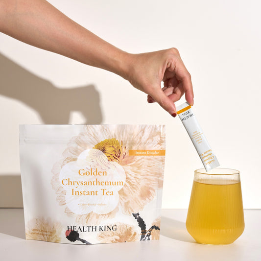 Hand holding a Health King Golden Chrysanthemum Instant Tea package with a glass of tea on a light background