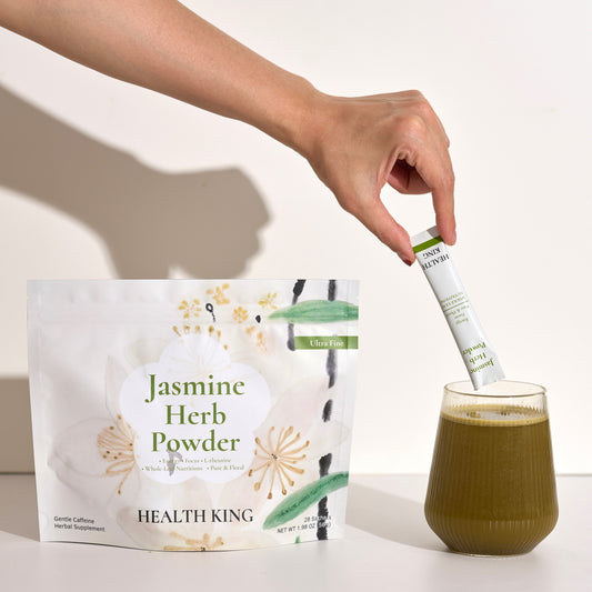 Hand holding a Jasmine Herb Powder package with a glass of green tea on a white background