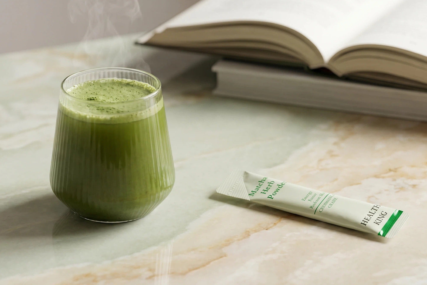Green smoothie in a glass with a package of matcha powder on a marble surface, next to an open book.