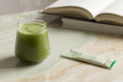 Green smoothie in a glass with a package of matcha powder on a marble surface, next to an open book.
